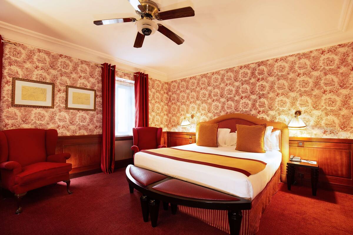 Habitación de hotel con una cama grande, un sillón rojo, una banca, un ventilador de techo y un papel tapiz estampado.