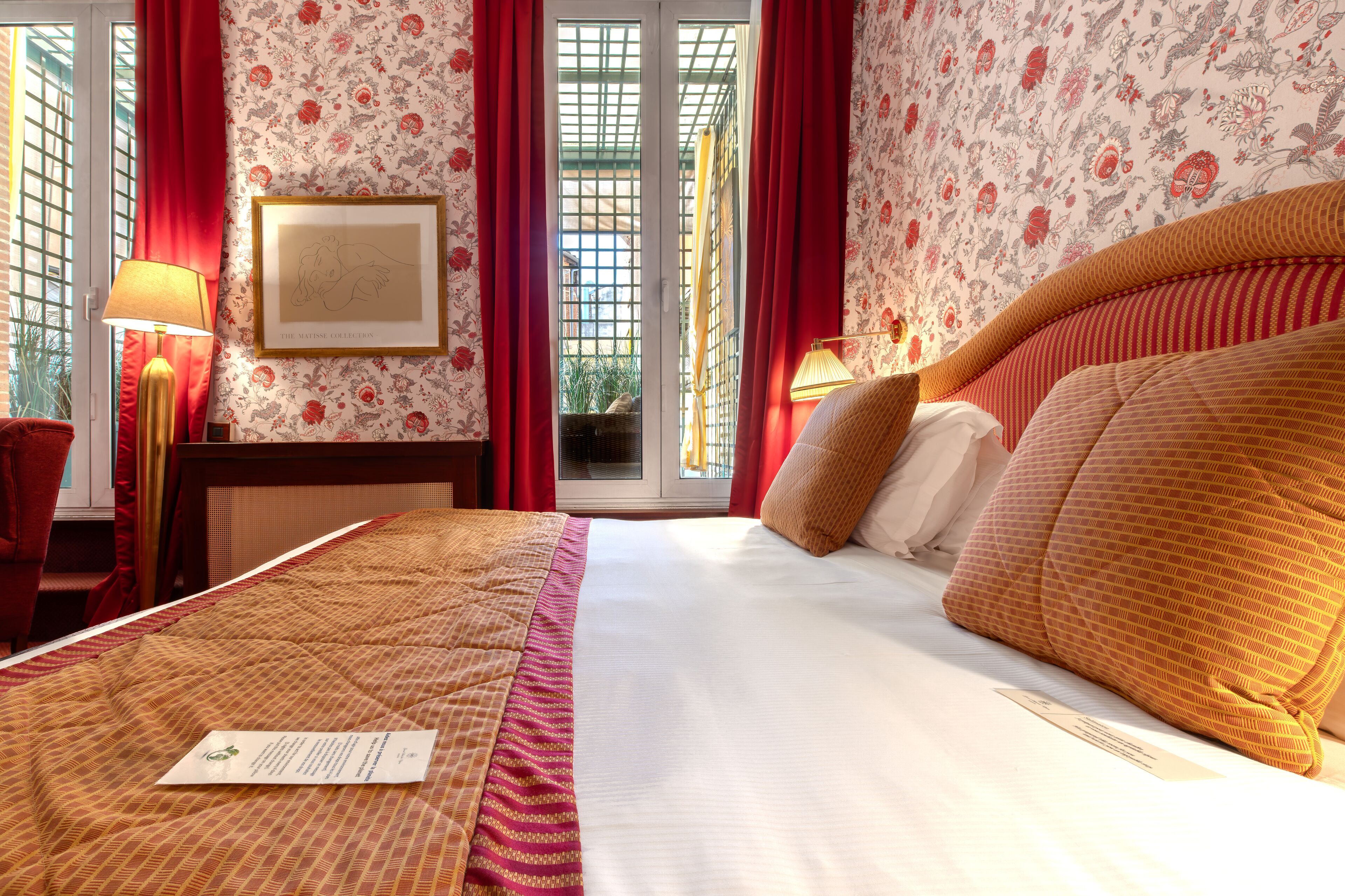 Una habitación de hotel con una cama grande, cortinas rojas, papel tapiz floral y un cuadro en la pared.