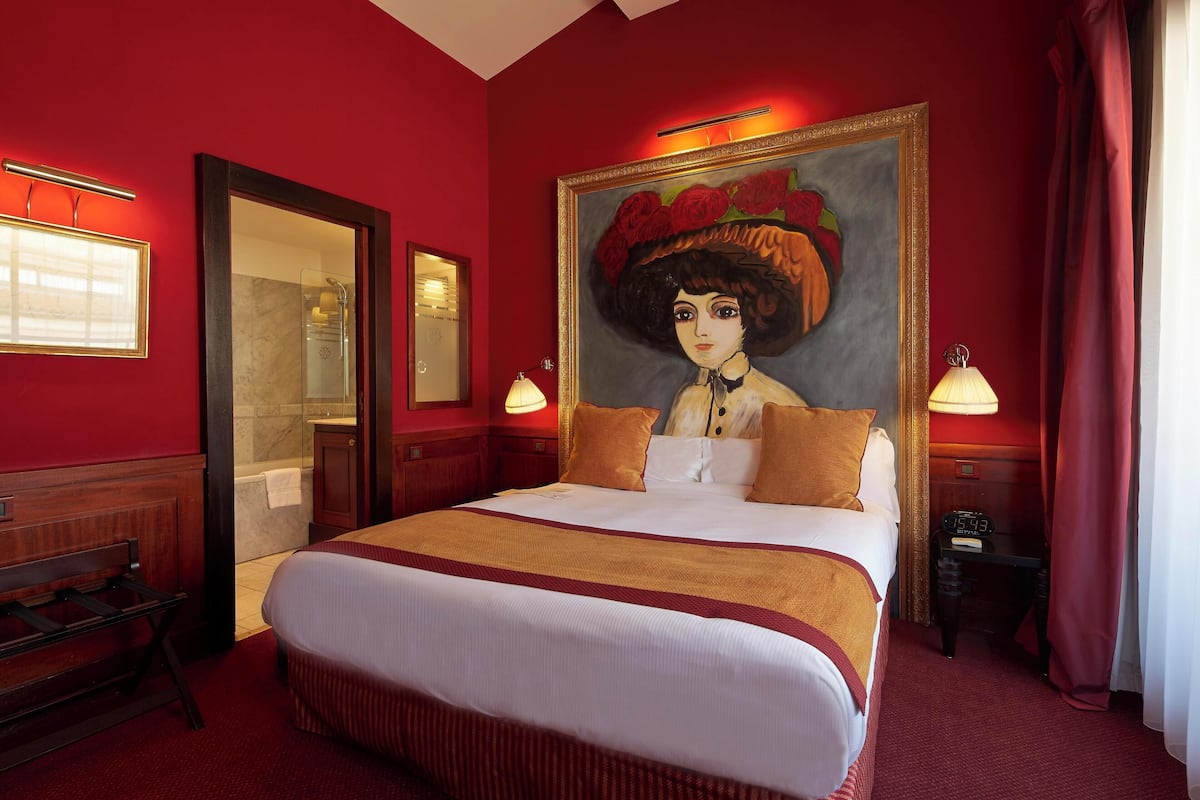 Una habitación de hotel con una cama grande, un cuadro en la pared, un espejo y una mesita de noche con una lámpara.