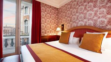 A hotel room with a large bed, a window with a view of buildings, and a patterned wallpaper.
