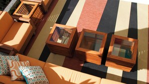 Un área de estar al aire libre con luz solar, suelo a rayas, un sofá y dos mesas cuadradas con tapas de vidrio.