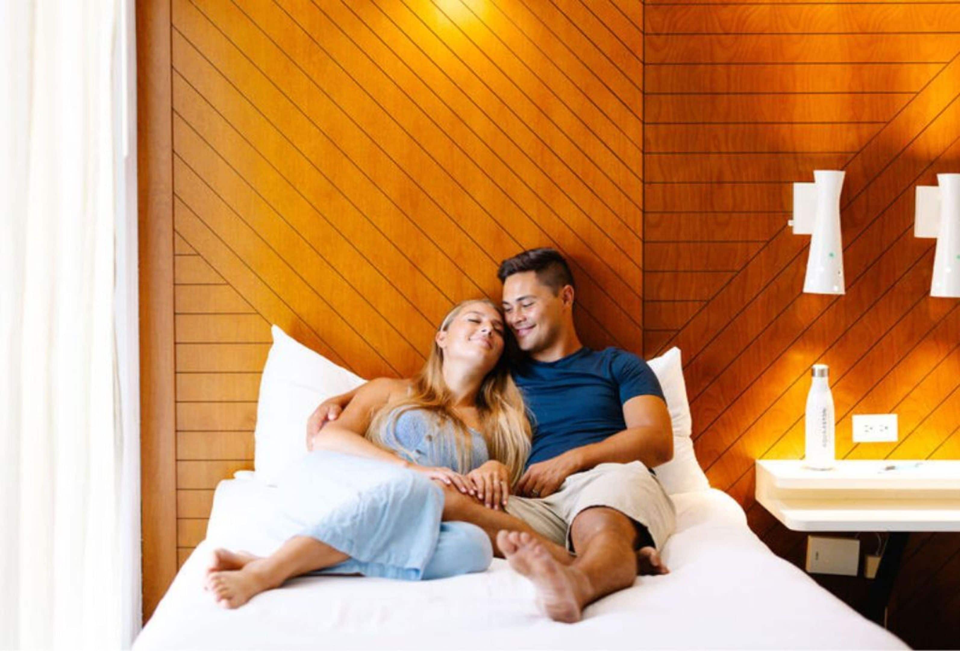 A couple on a bed in a room with wooden paneling and a bedside table with a bottle.