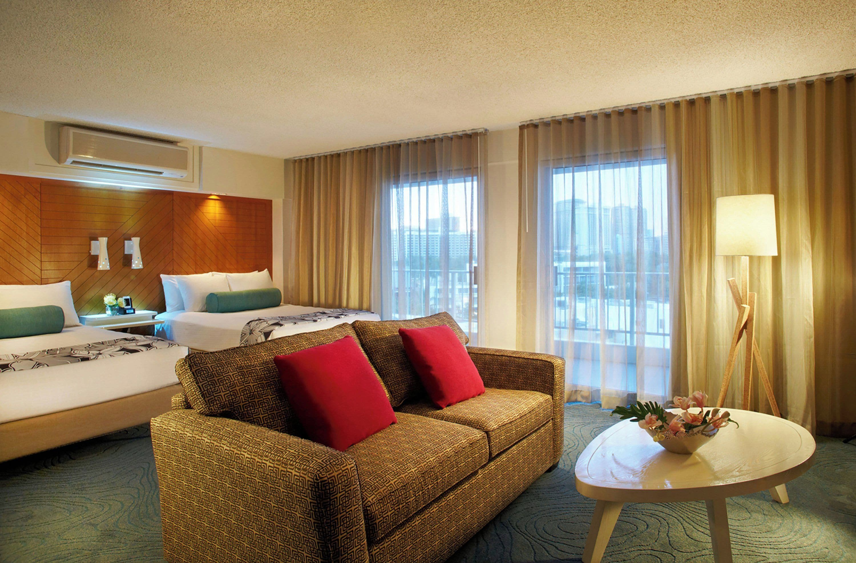 Habitación de hotel con cama, sofá, una mesa con lámpara y vistas a la ciudad a través de amplias ventanas.