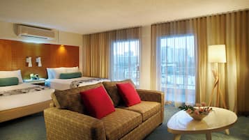 Habitación de hotel con cama, sofá, una mesa con lámpara y vistas a la ciudad a través de amplias ventanas.