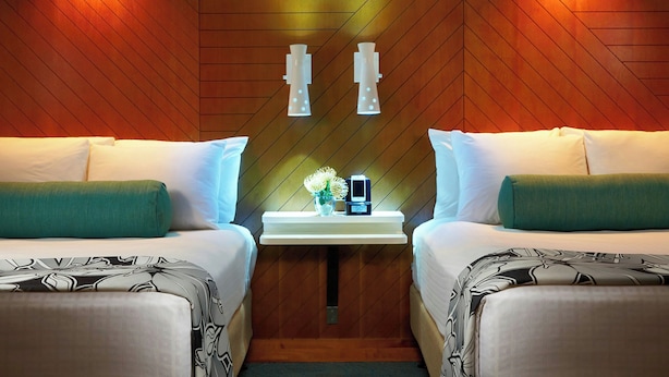 A hotel room with two beds, a nightstand with a clock and a vase of flowers, and wooden paneling on the walls.