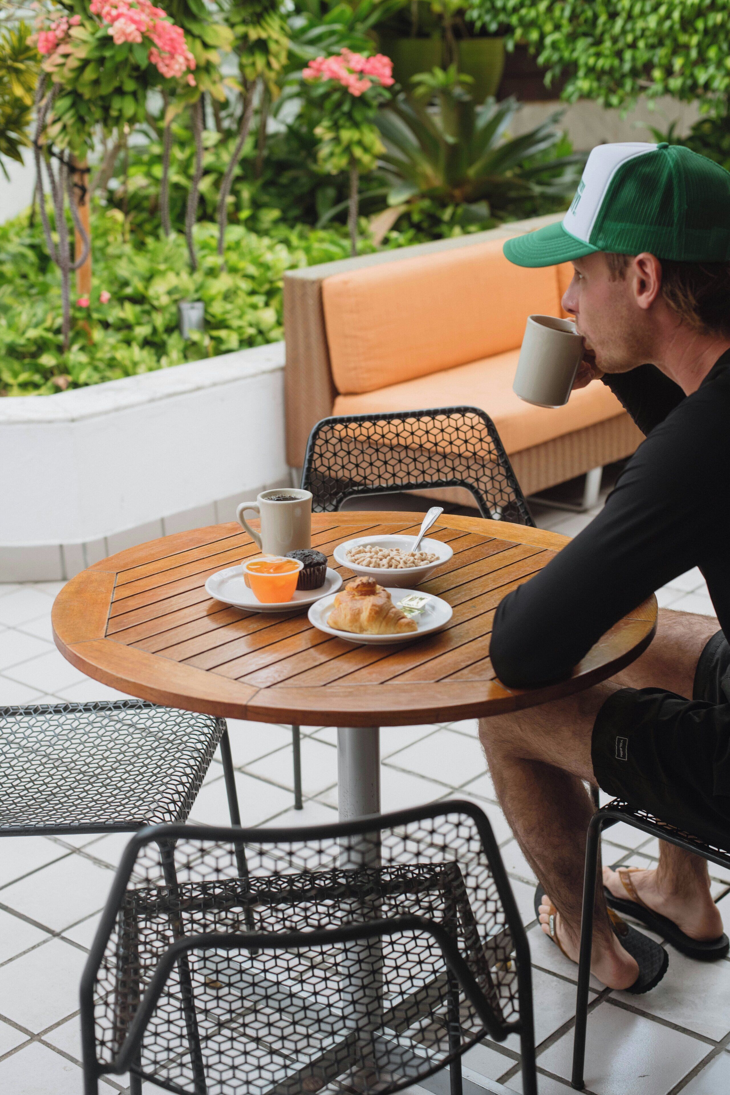 A person seated at a round wooden table with a cup, a bowl of cereal, and a muffin. There is a metal chair in front of the table, and a mesh chair to the left.