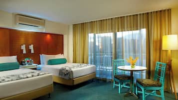 Ein Hotelzimmer mit zwei Betten, einem kleinen Tisch mit Stühlen, einer Lampe und Blick auf die Stadt durch leichte Vorhänge.