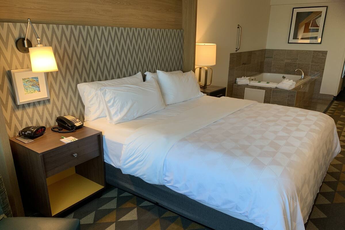 Ein Hotelzimmer mit einem großen Bett, einem Nachttisch, einer Lampe, einem Telefon am Bett, einem Badezimmer mit einer Badewanne und einem Handtuchhalter.