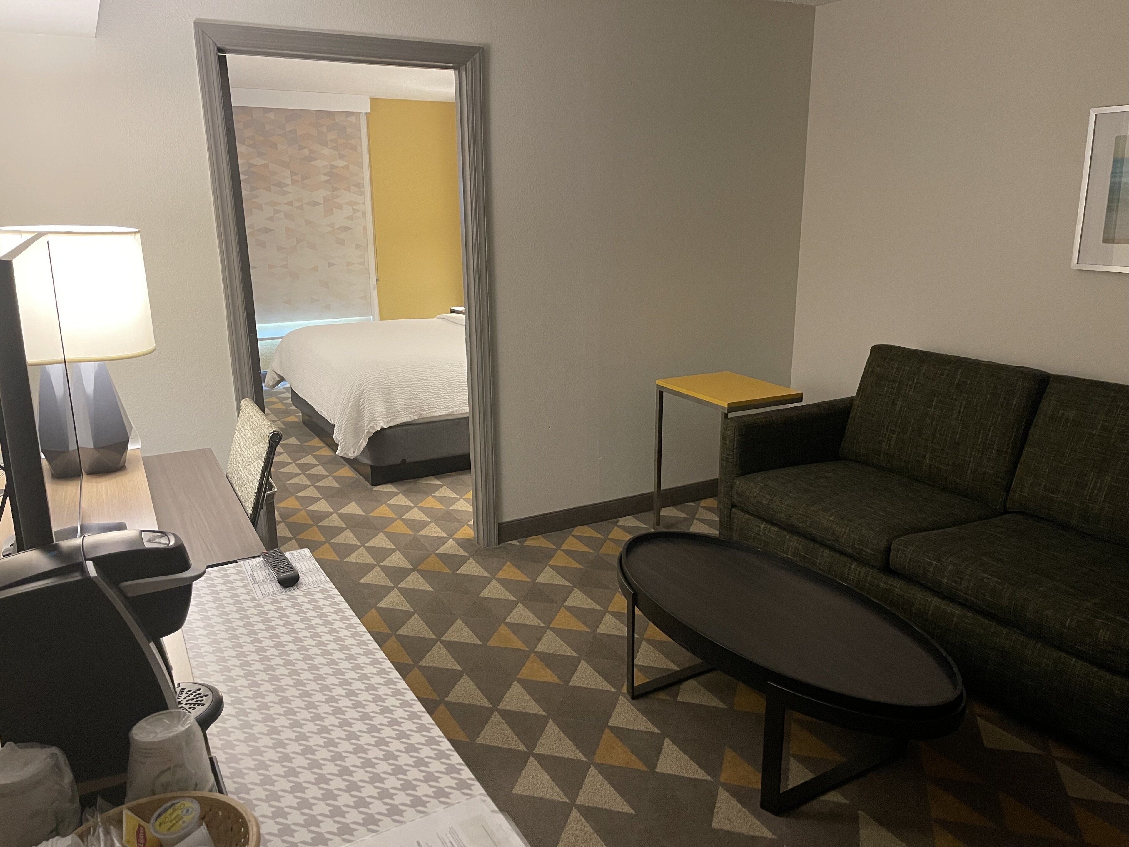 Quarto de hotel com cama, sofá, mesa pequena, luminária e escrivaninha com computador.