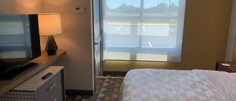 Habitación de hotel con cama, televisor, escritorio con lámpara, cómoda y ventana con vista a un estacionamiento.
