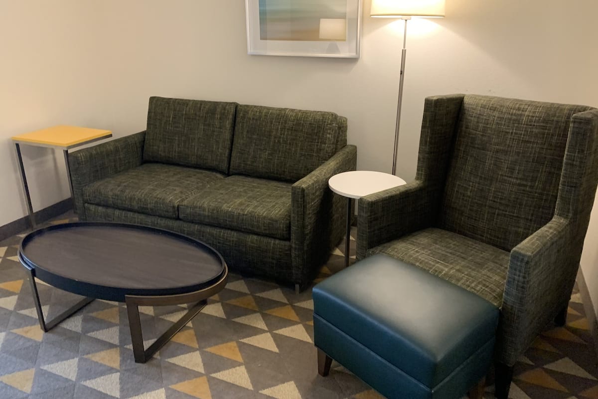 Ein Zimmer mit einem grünen Sofa, einem blauen Hocker, einem kleinen weißen Tisch und einer Stehlampe.