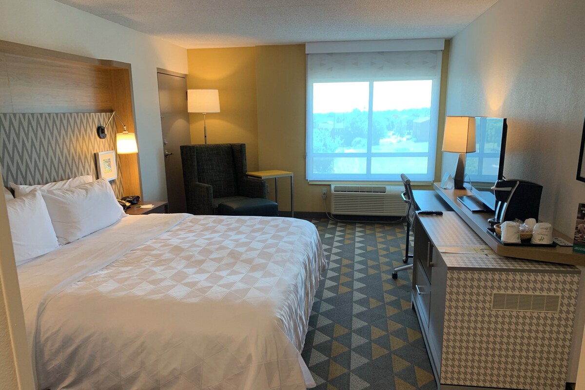 Une chambre d’hôtel avec un grand lit, un bureau, une chaise, une lampe de bureau, une fenêtre et un téléviseur fixé au mur.