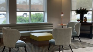 Un salon moderne avec un canapé d’angle, deux fauteuils, une petite table et un lampadaire. De grandes fenêtres offrent une vue sur l’extérieur.