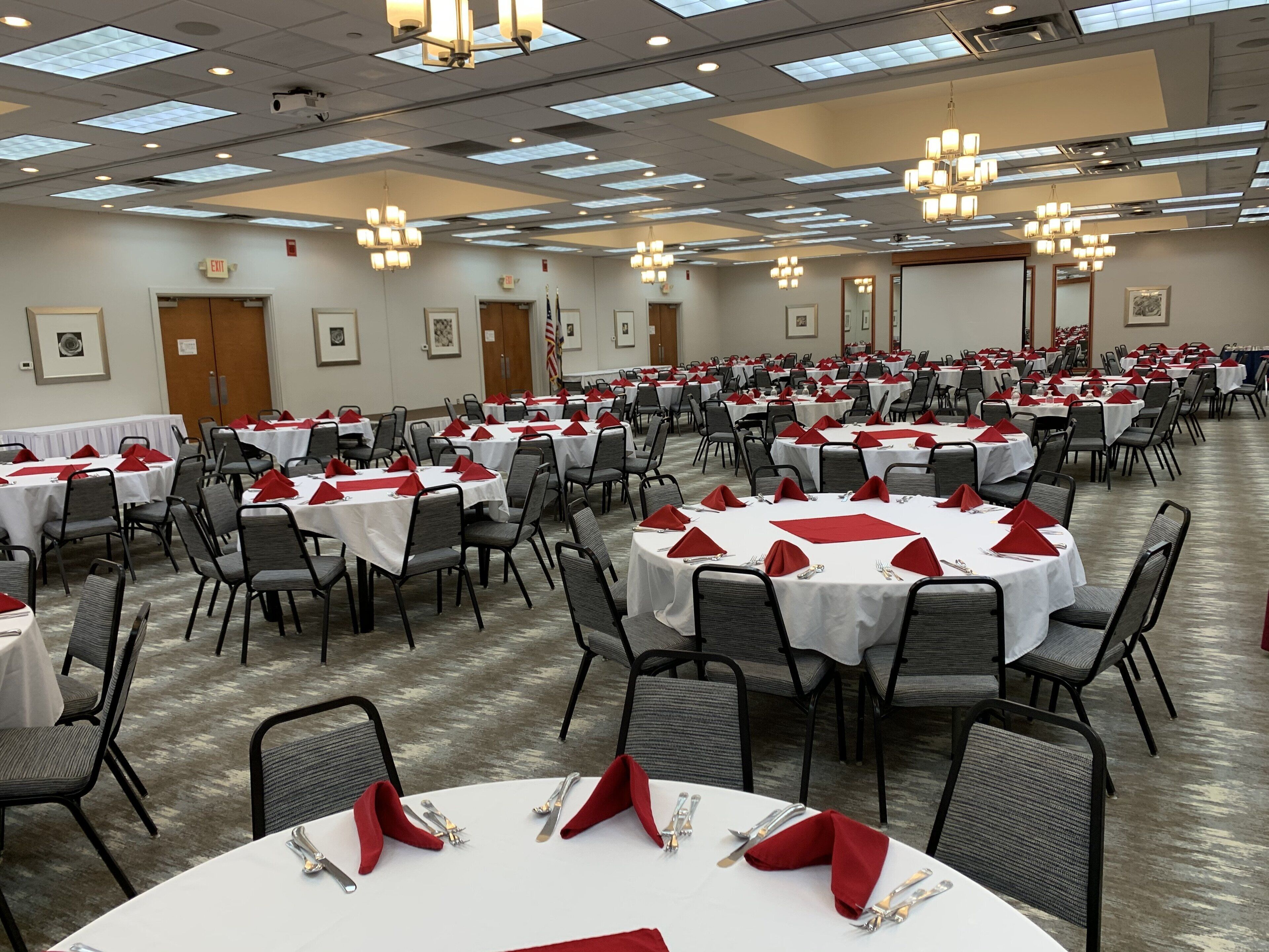 A large banquet hall with multiple round tables, each set with white tablecloths, red napkins, and silverware.