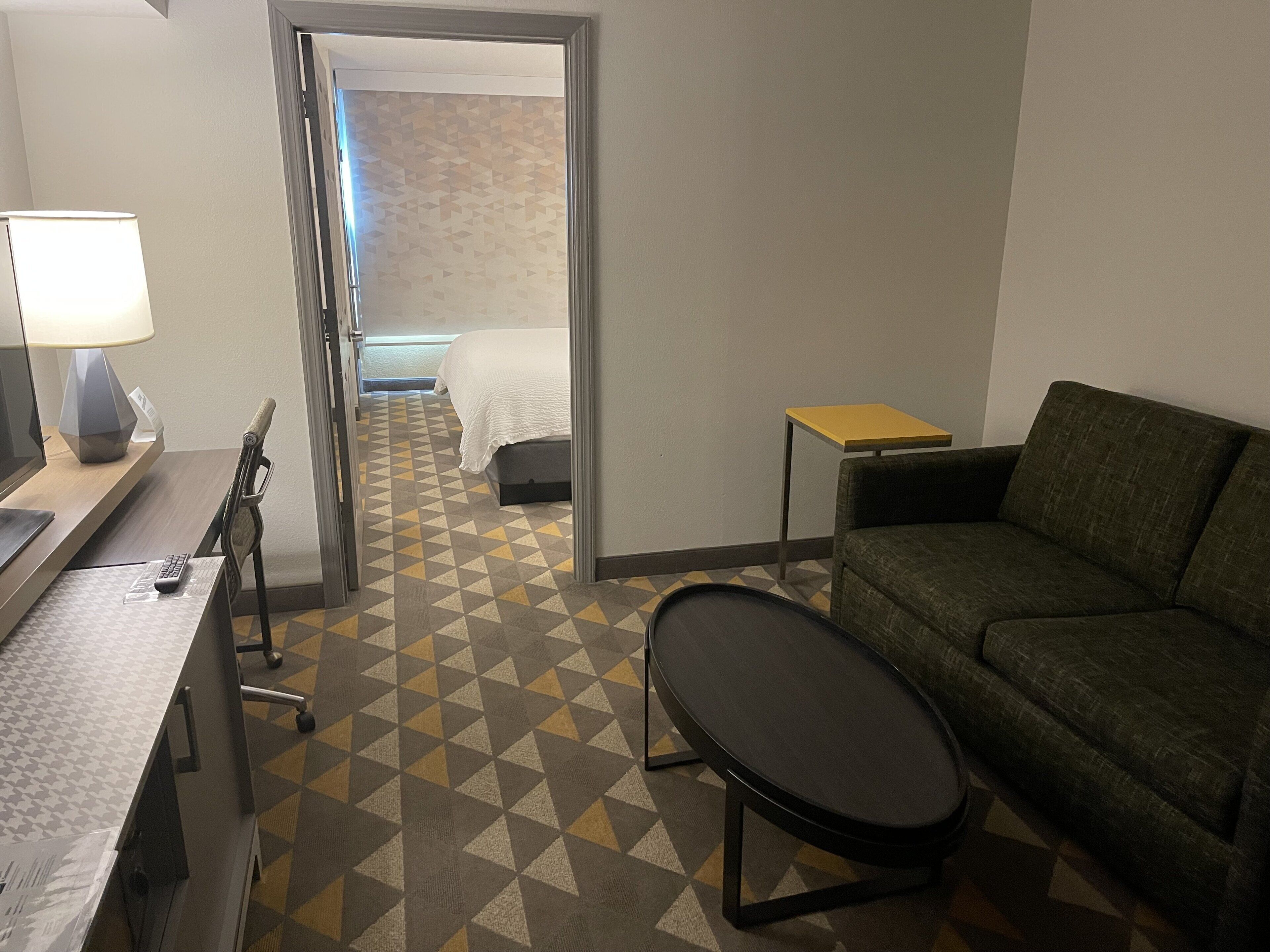 A hotel room with a bed, a sofa, a small table, a lamp, and a desk with a computer and remote control.