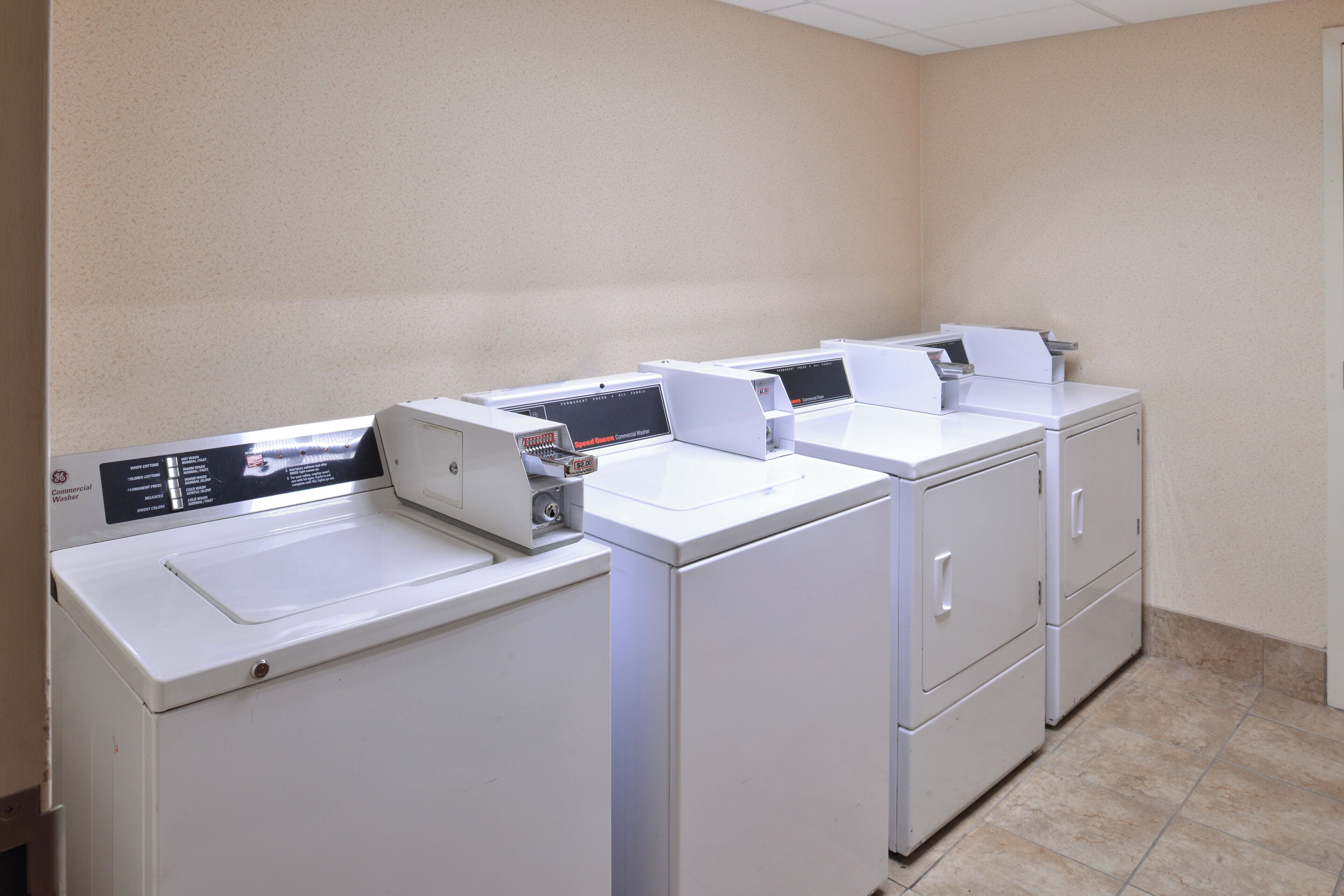 A row of white commercial washers in a room with beige walls and a tiled floor.