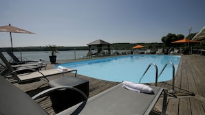 Un espace aménagé au bord de la piscine, avec des chaises longues, des parasols et une terrasse en bois donnant sur une étendue d’eau.