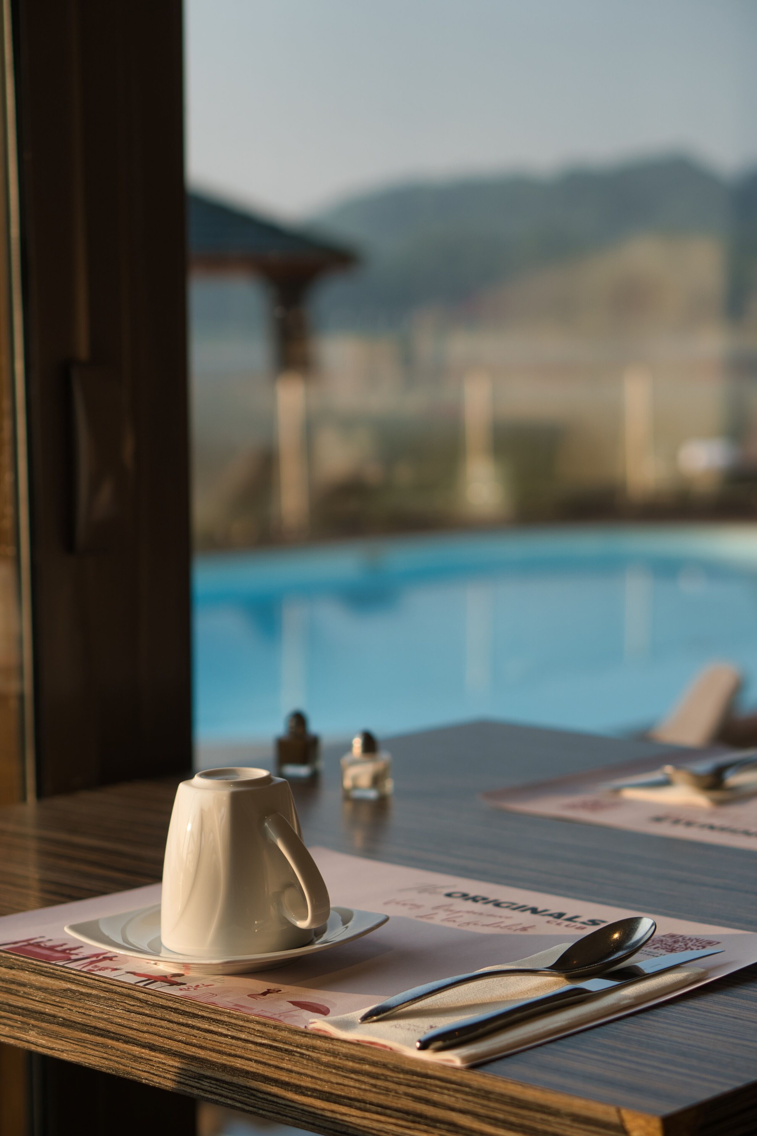 A table with a white cup and saucer, a spoon, and a knife on a placemat with a menu. A pool is visible in the background.
