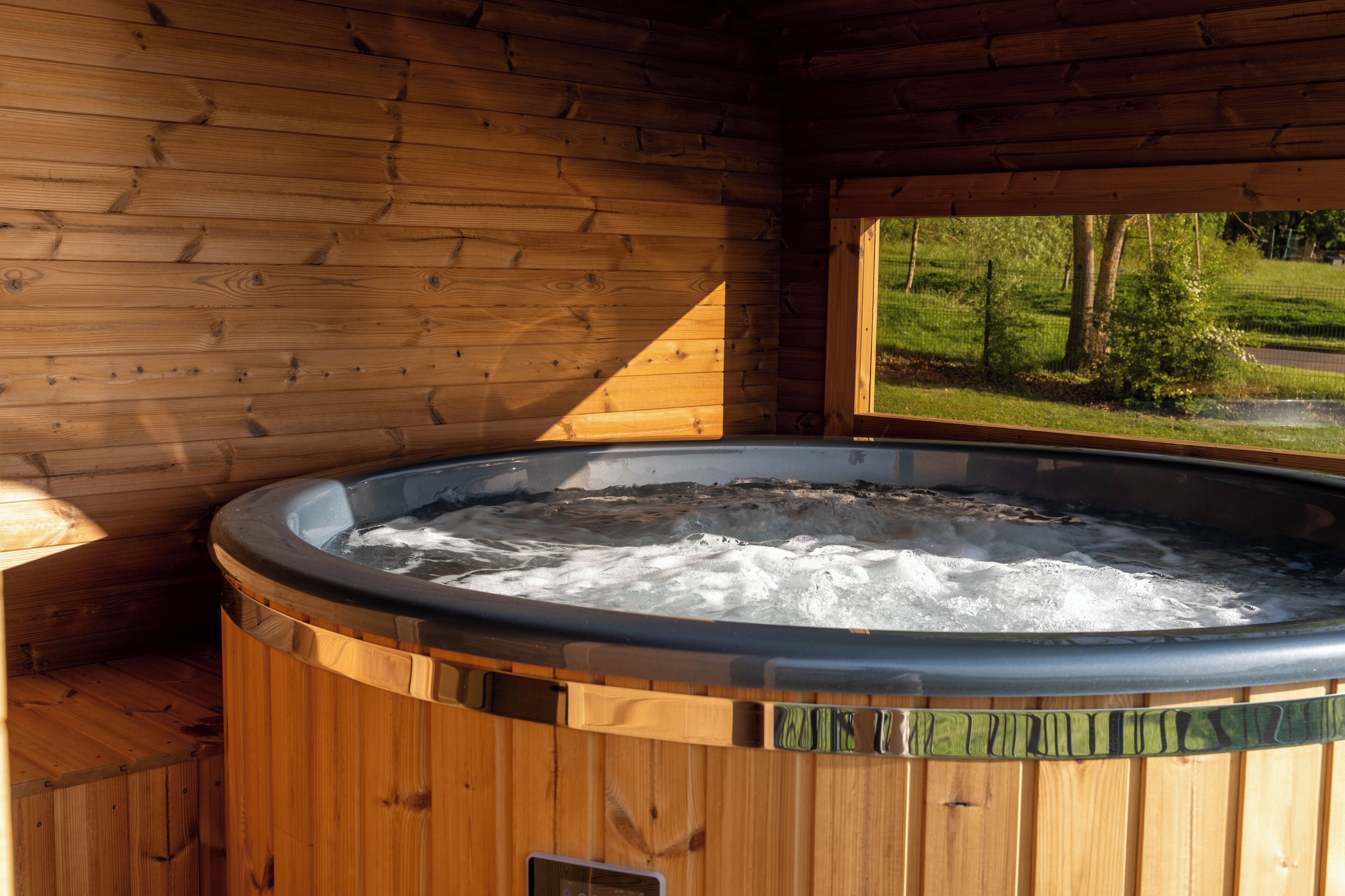 A wooden cabin interior with a hot tub, wooden walls, and a window overlooking a green landscape.