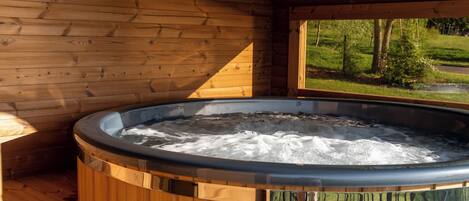 Interior de una cabaña de madera con jacuzzi, paredes de madera y ventana con vistas a un paisaje verde.