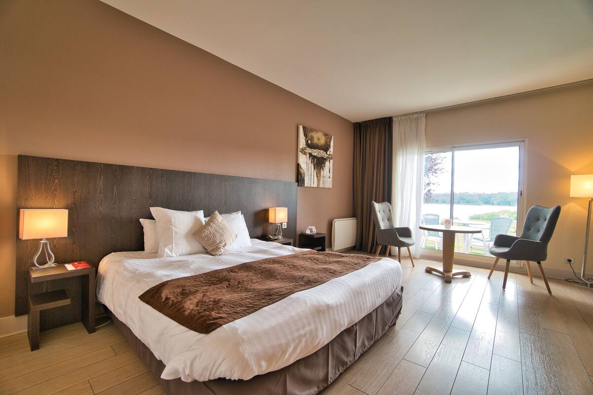 Een hotelkamer met een groot bed, een nachtkastje, een tafel met stoelen en uitzicht op de omgeving.