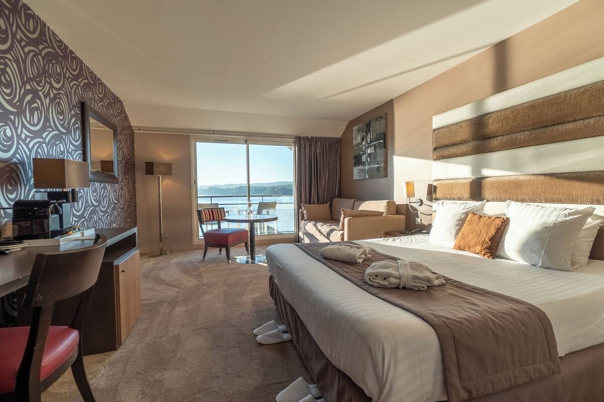 Een hotelkamer met een groot bed, een bureau met stoel, een eettafel met stoelen en uitzicht op zee.