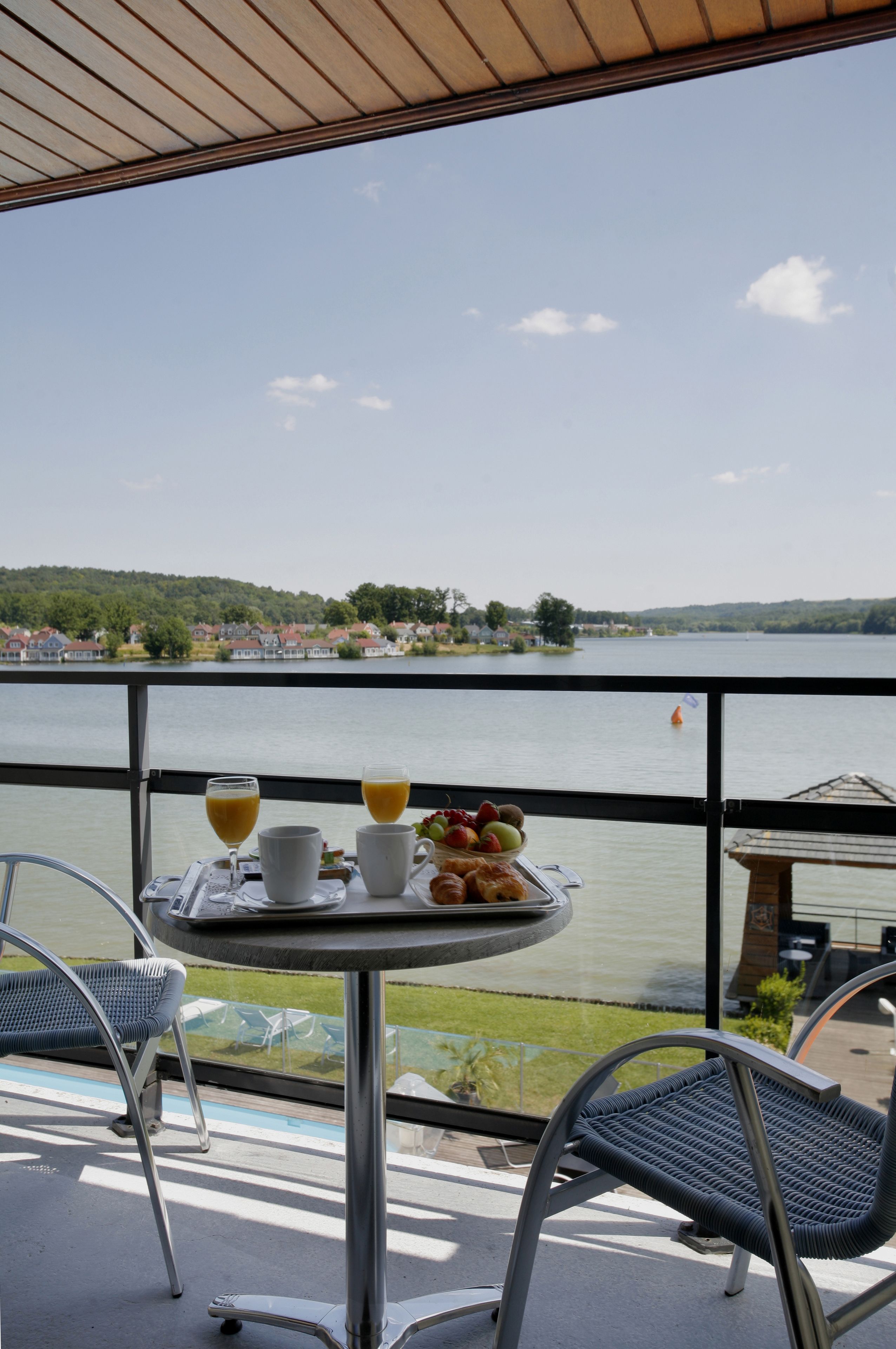 Un balcón con una mesa dispuesta para el desayuno, con vista a un lago y casas al fondo.