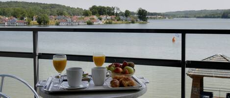 Un balcón con una mesa dispuesta para el desayuno, con vista a un lago y casas al fondo.