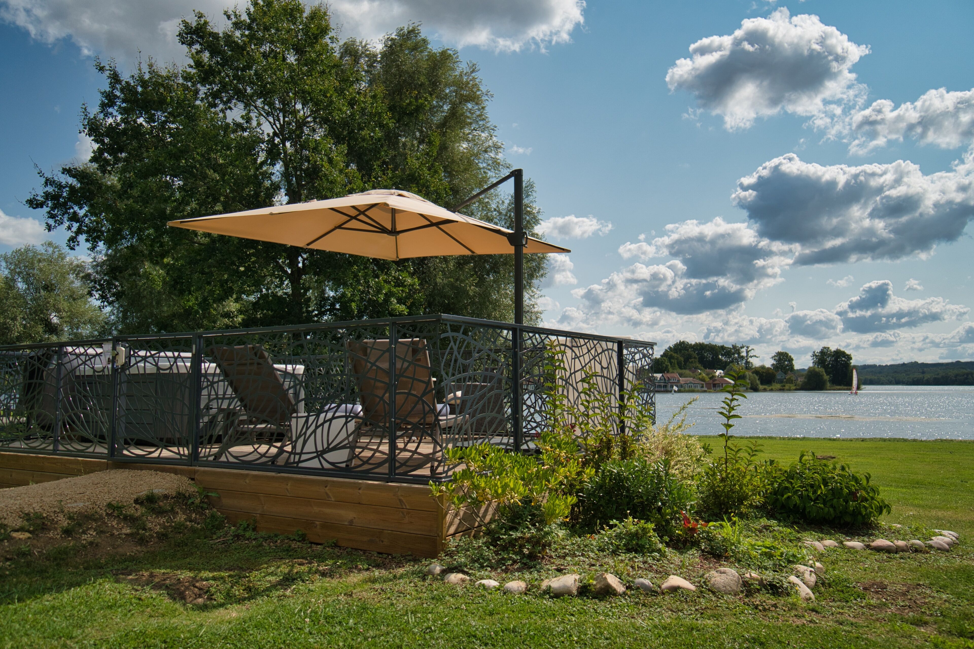 Una terraza con mobiliario exterior y un paraguas grande, con vista a un lago y un velero a lo lejos.