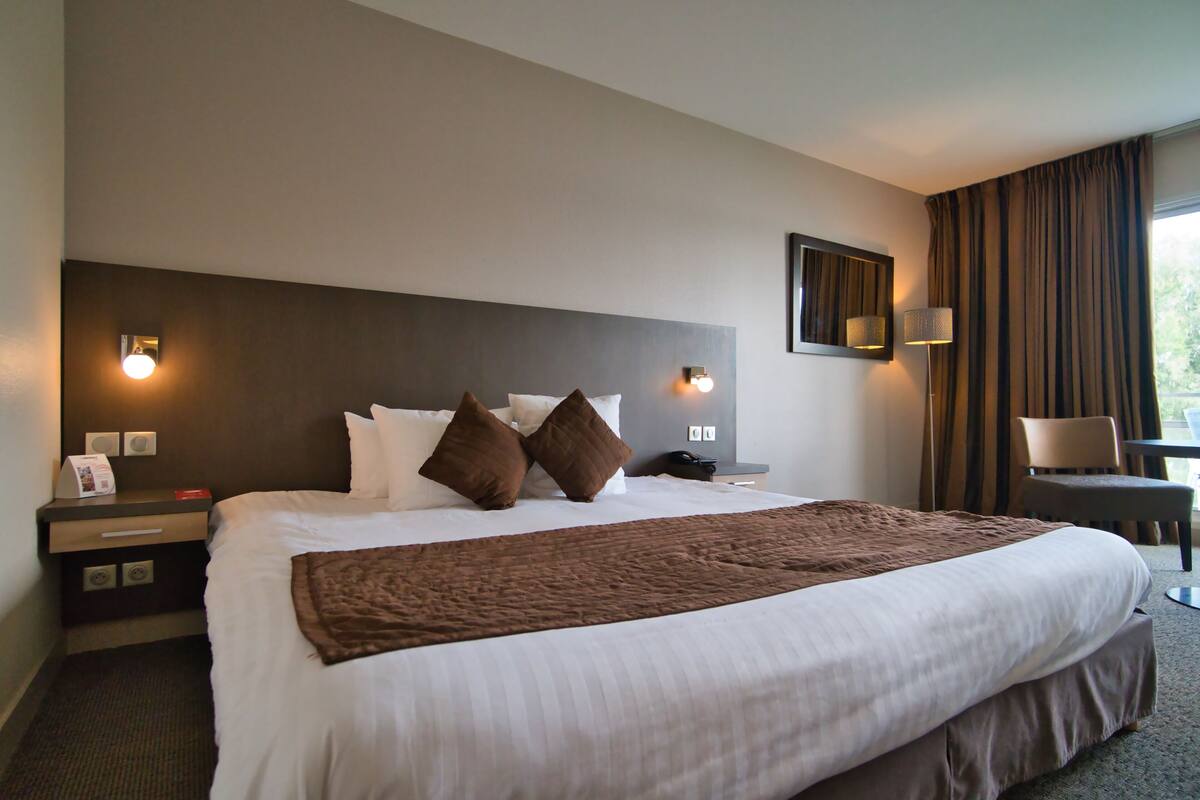 Hotelkamer met een groot bed, twee nachtkastjes, een bureau, een stoel en een raam met gordijnen.