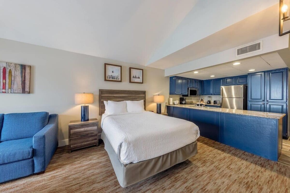 Een hotelkamer met een bed, een blauwe fauteuil, een klein bureau en een kitchenette met blauwe kastjes en een roestvrijstalen koelkast.