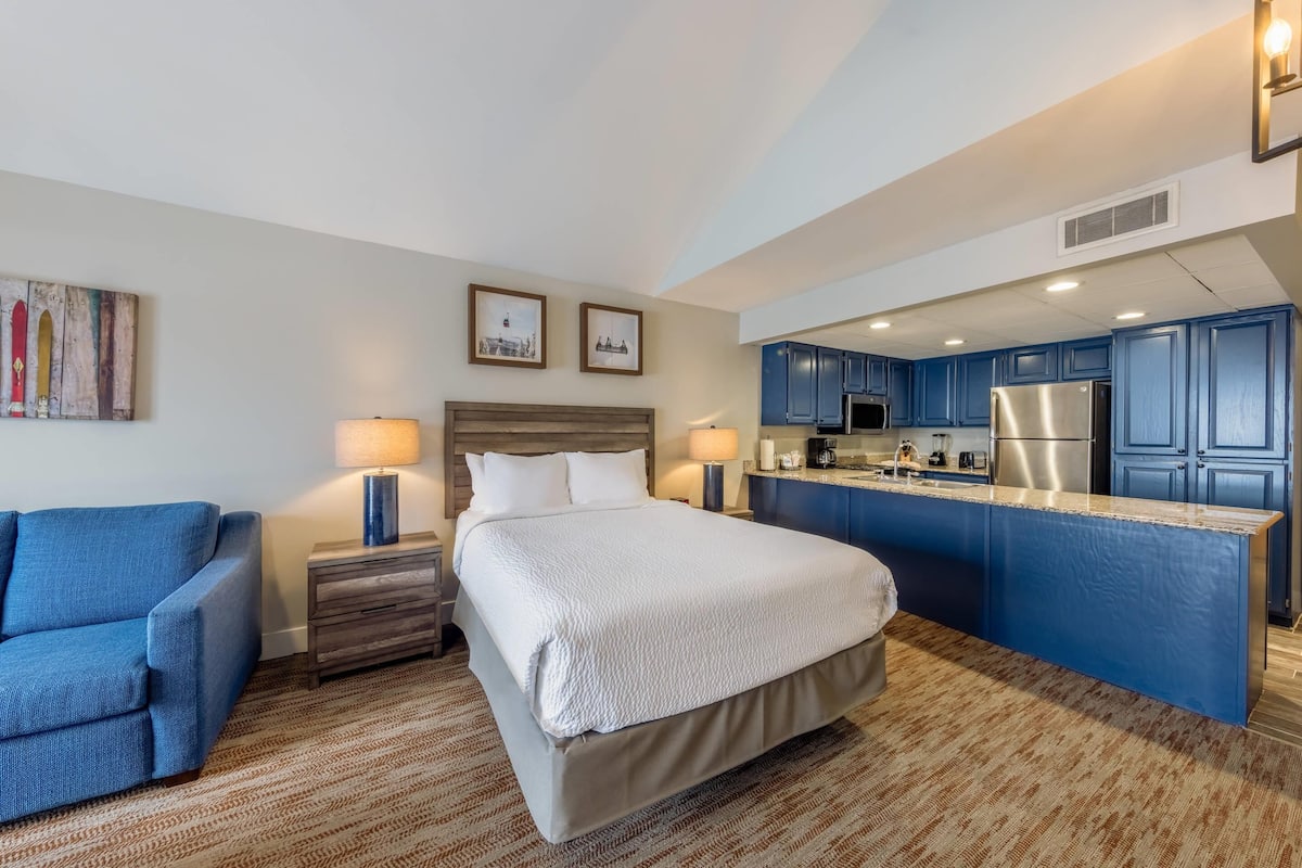 Een hotelkamer met een bed, een blauwe fauteuil, een klein bureau en een kitchenette met blauwe kastjes en een roestvrijstalen koelkast.