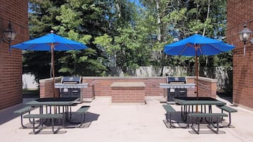 An outdoor patio with two barbecue grills, two picnic tables, and two blue umbrellas.