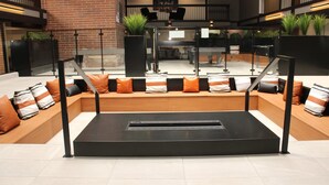 Een moderne lounge met een centrale zwarte tafel, comfortabele zithoek met oranje en gestreepte kussens, en potplanten.