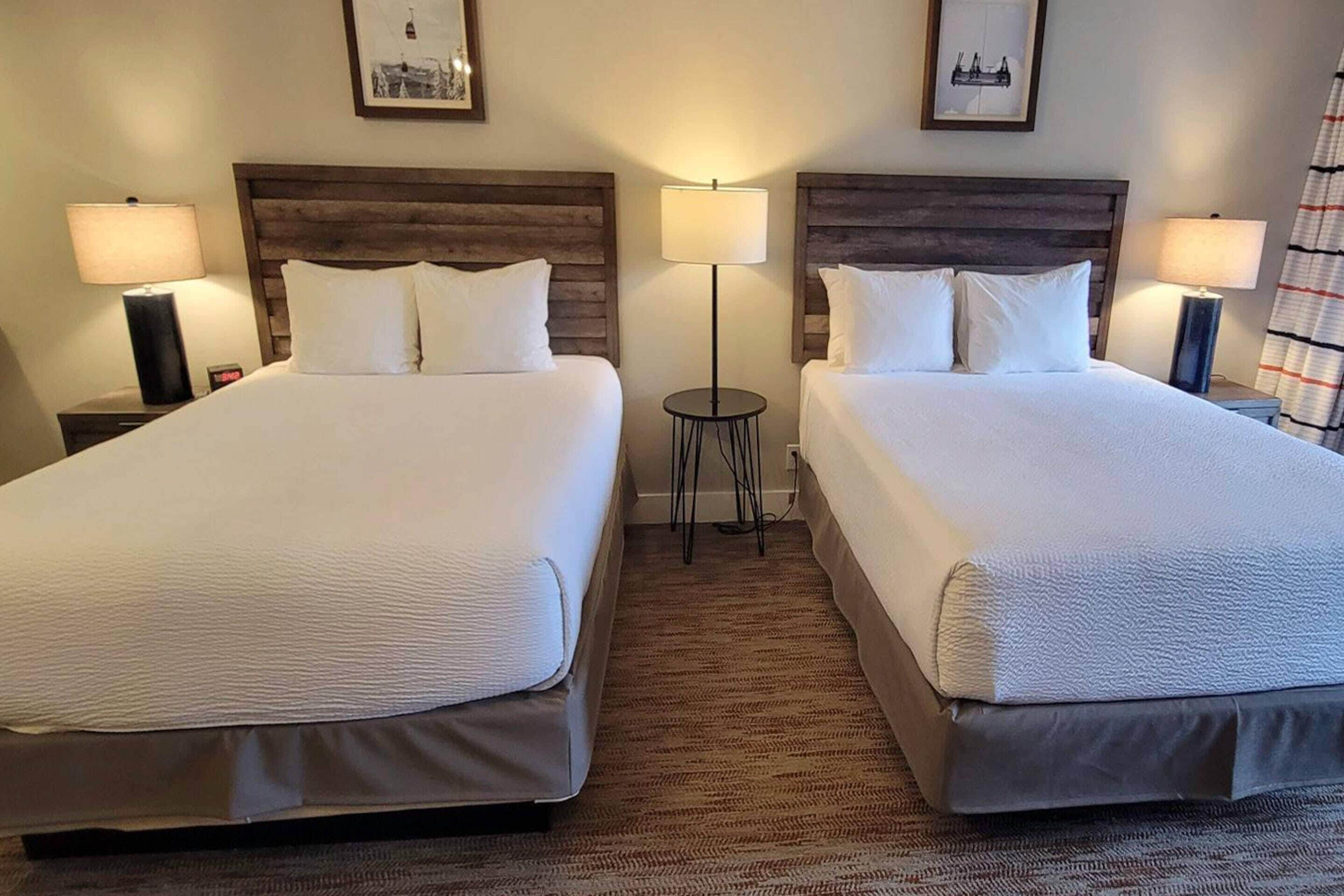 A hotel room with two beds, each with a wooden headboard, white bedding, and bedside lamps.