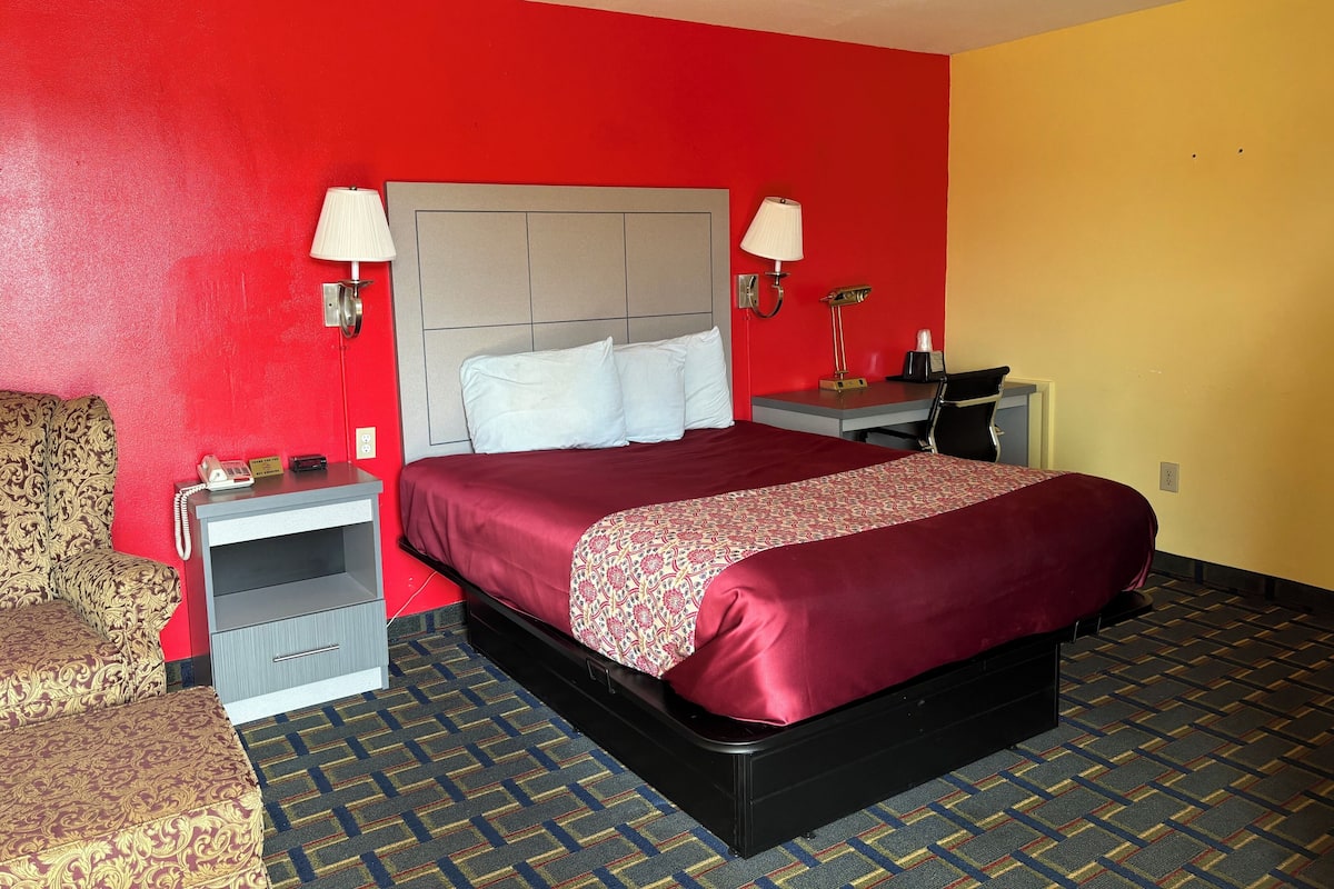 A hotel room with a red and yellow wall, a bed with a patterned bedspread, two bedside lamps, a desk with a lamp, a chair, and a patterned carpet.