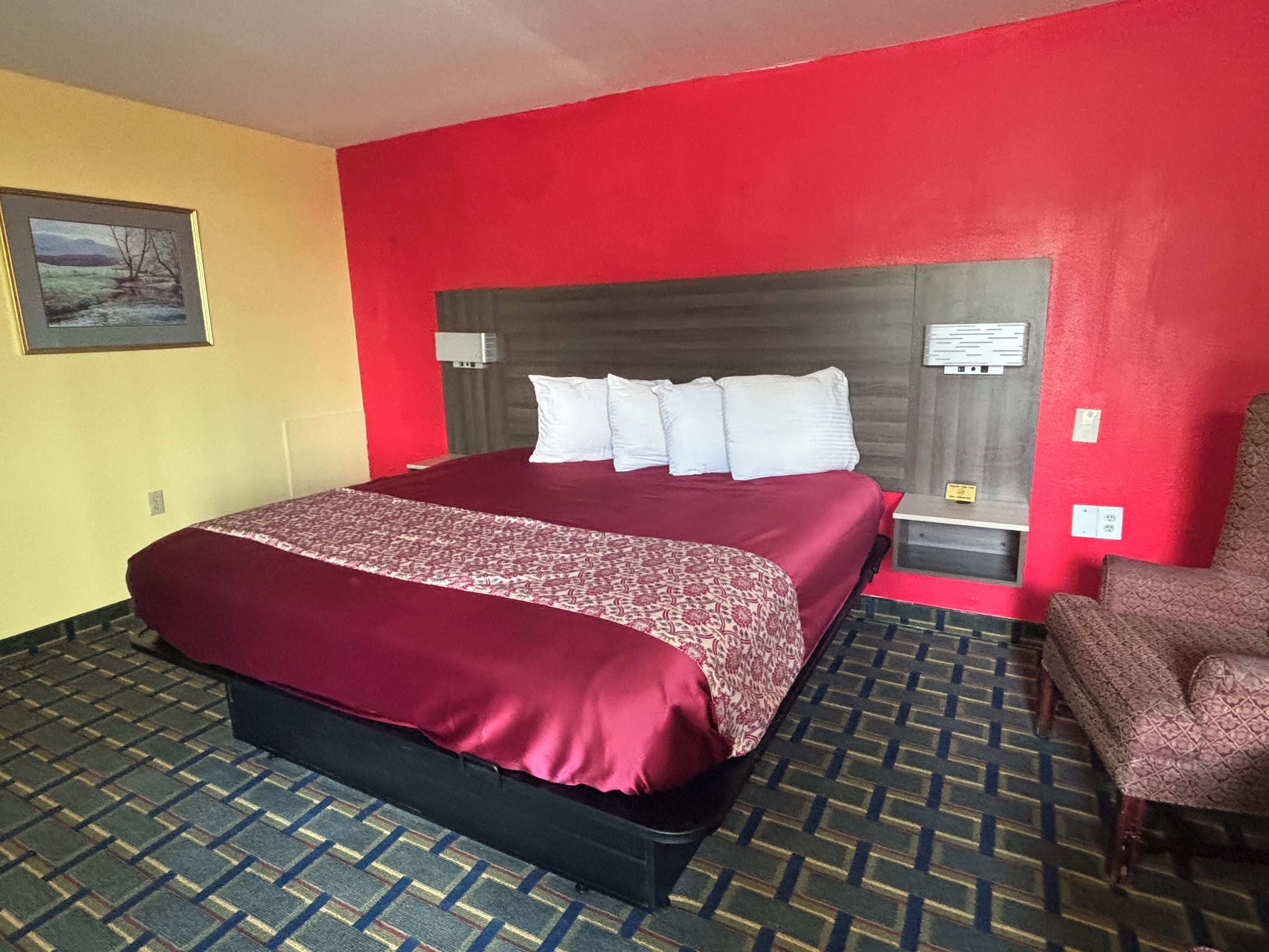 A hotel room with a large bed, a red accent wall, a patterned carpet, and a chair.