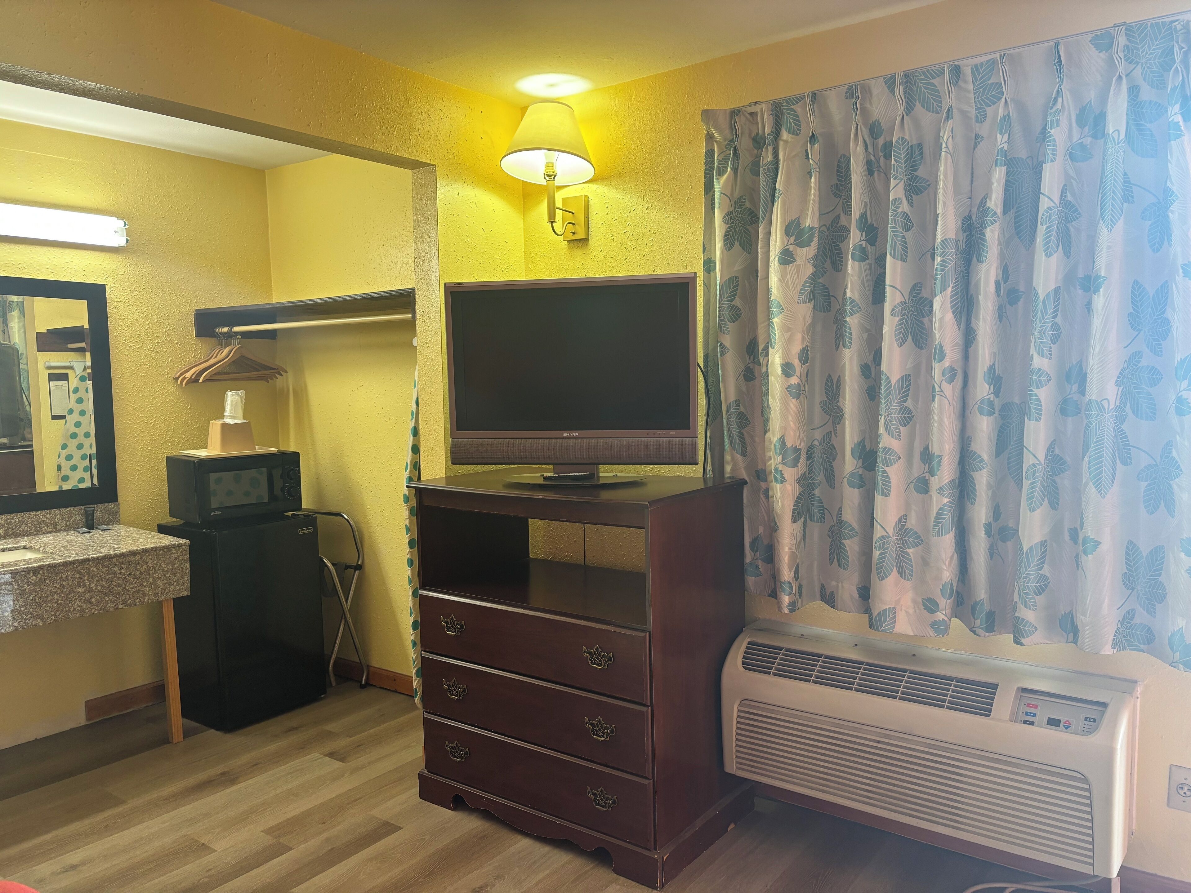 A hotel room with a wooden dresser, a flat-screen TV, a microwave, a heater, and a mirror.