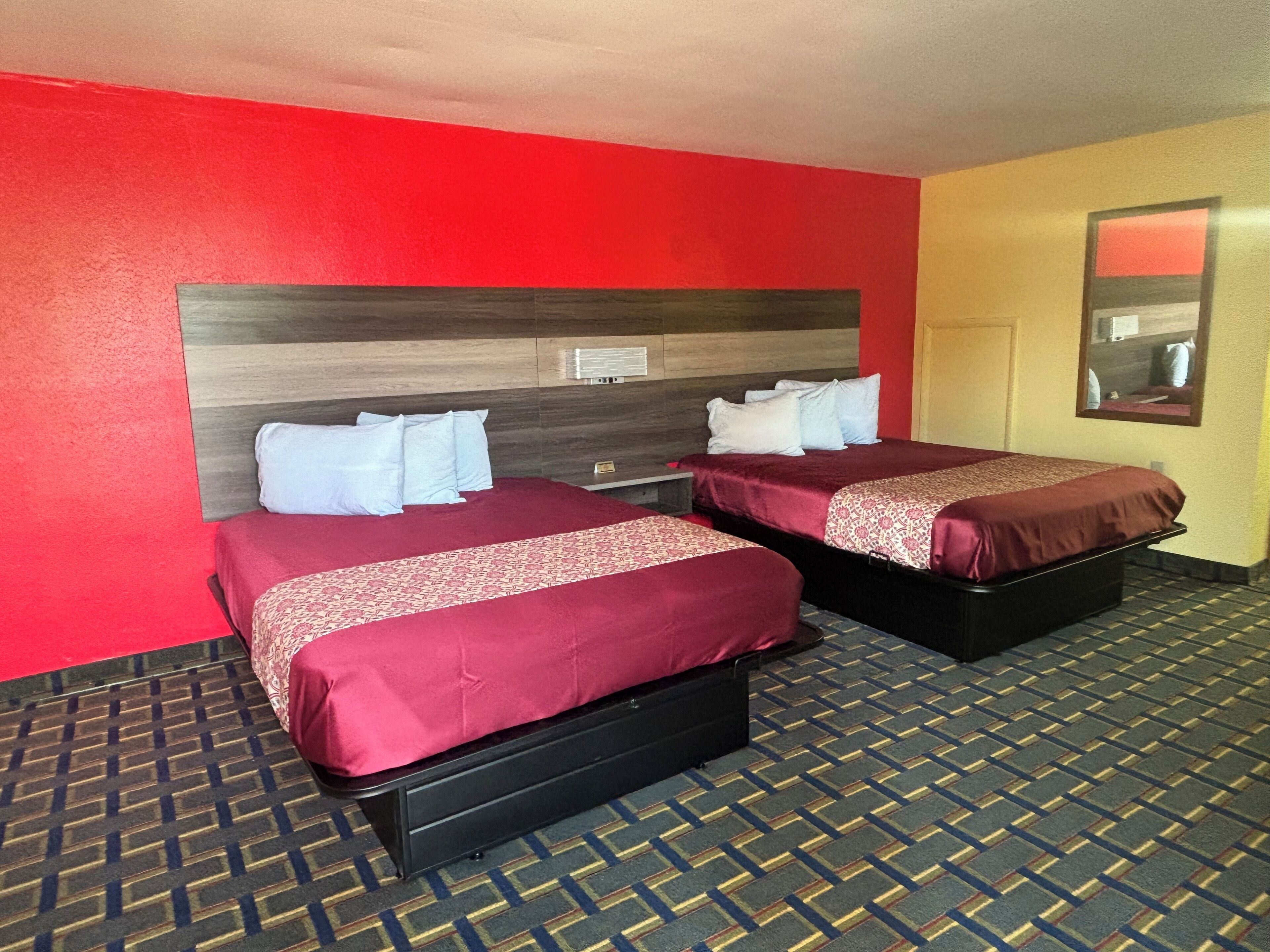A hotel room with two beds, each with a red bedspread and white pillows. The room has a red wall and a patterned carpet.