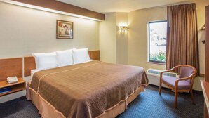 Una habitación de hotel con una cama grande, una silla, una mesita de noche, una lámpara de pared y un ventanal con cortinas.