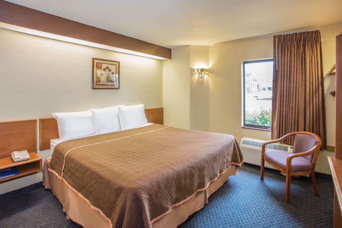 Une chambre d’hôtel avec un grand lit, une chaise, une table de chevet, une lampe fixée au mur et une fenêtre avec des rideaux.