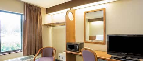 A hotel room with a bed, two chairs, a desk with a microwave, a television, and a window with curtains.