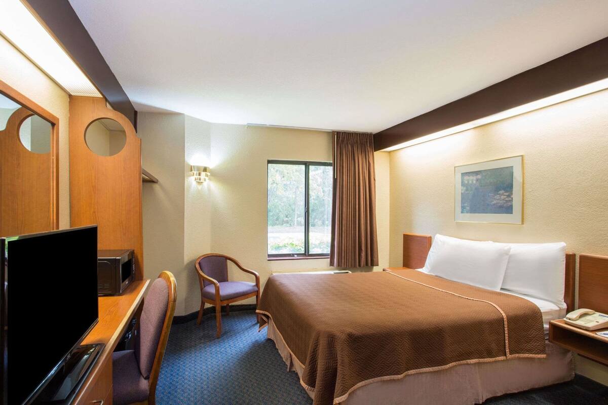 Une chambre d’hôtel avec un lit, un bureau avec une télévision, une chaise, une fenêtre avec des rideaux et un tableau au mur.