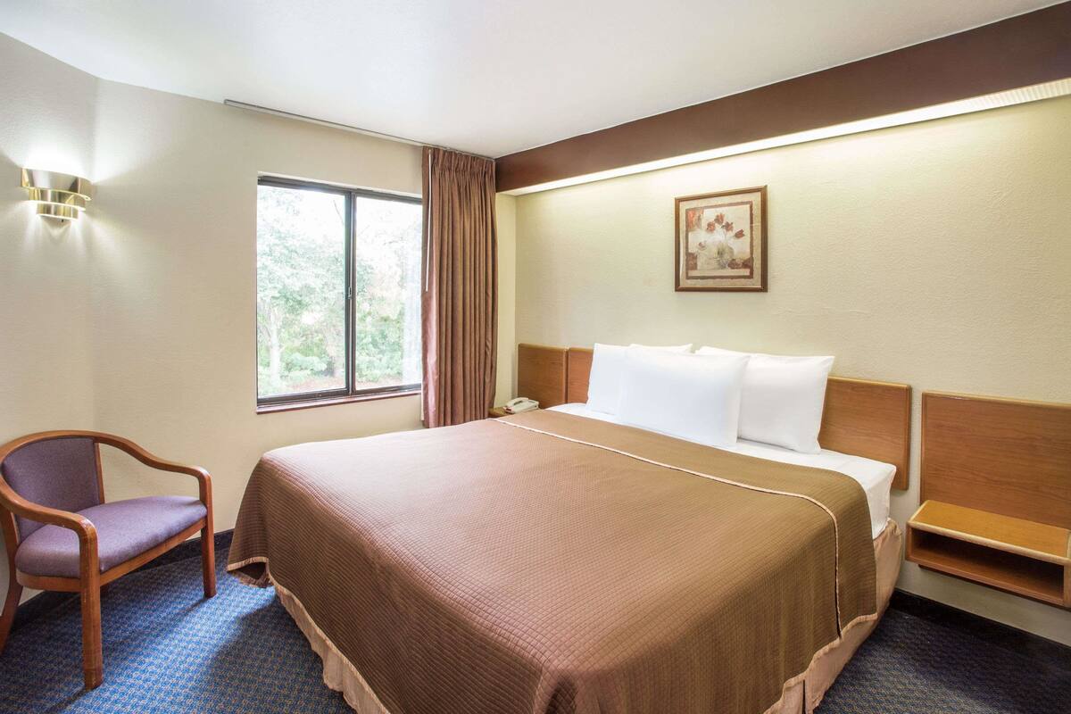Une chambre d’hôtel avec un grand lit, une chaise, une table de chevet, une fenêtre avec des rideaux et un luminaire fixé au mur.