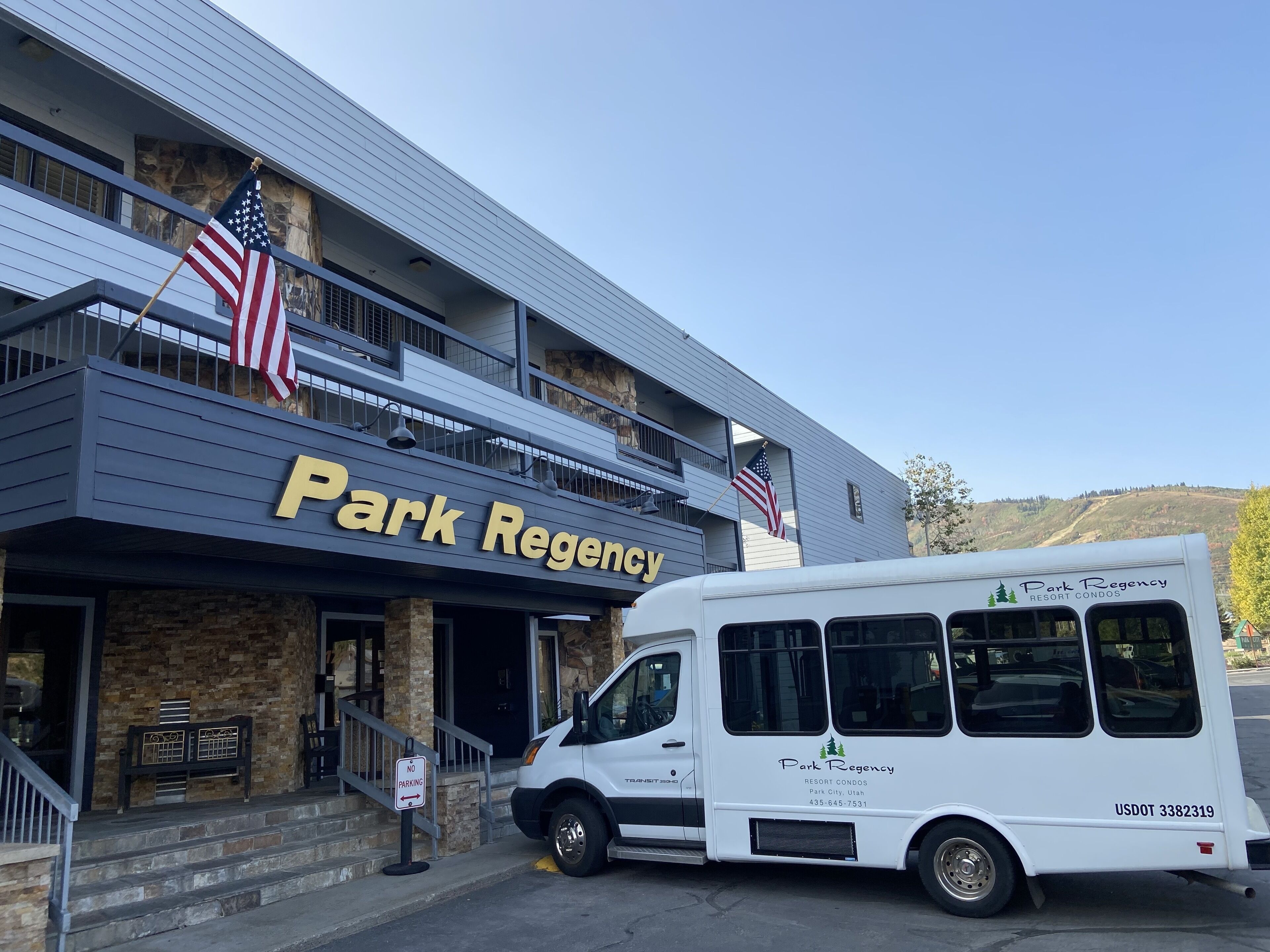 Um edifício com uma placa indicando "Park Regency" e uma bandeira americana. Uma van branca está estacionada na frente.
