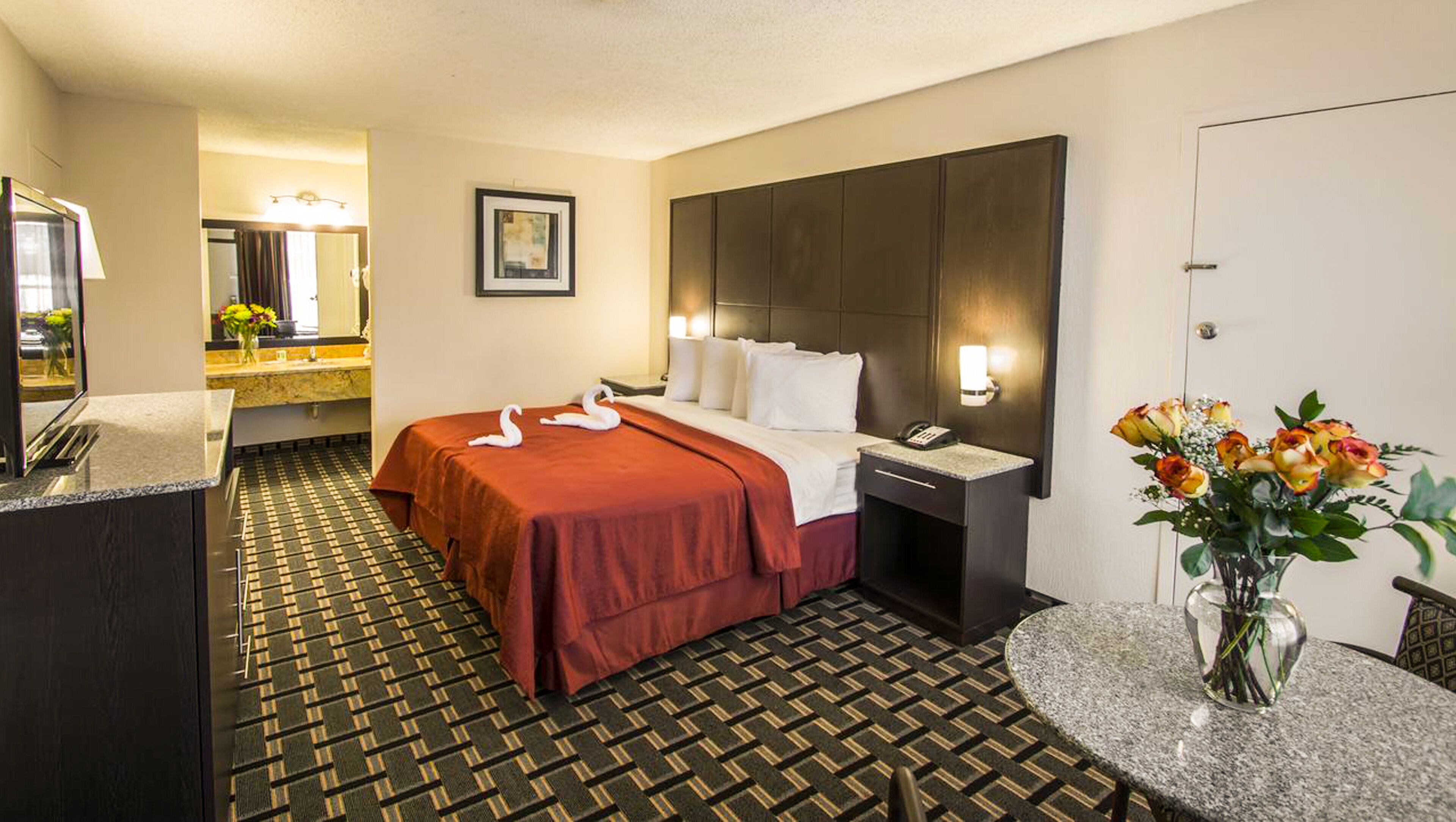 A hotel room with a bed, a bedside table with a lamp, a mirror, a vase with flowers, and a patterned carpet.