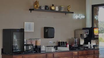 Una barra de cafetería con una máquina de café, un microondas, una caja registradora y una estantería con objetos decorativos.