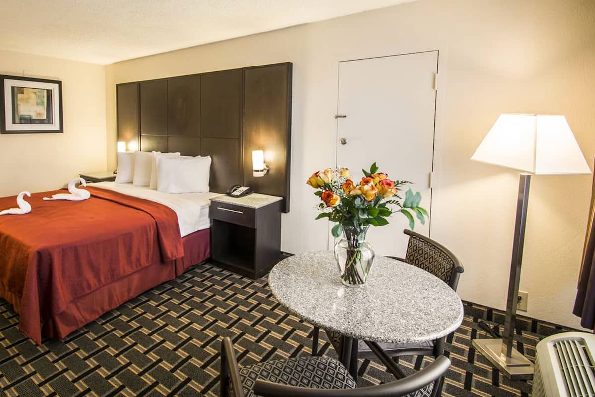 A hotel room with a bed, a desk, a chair, a table, a lamp, and a vase with flowers.