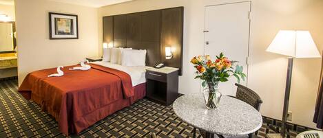 A hotel room with a bed, a desk, a chair, a table, a lamp, and a vase with flowers.