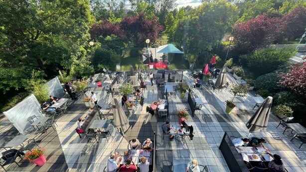 Een terras buiten met tafels en stoelen, omgeven door bomen en planten.